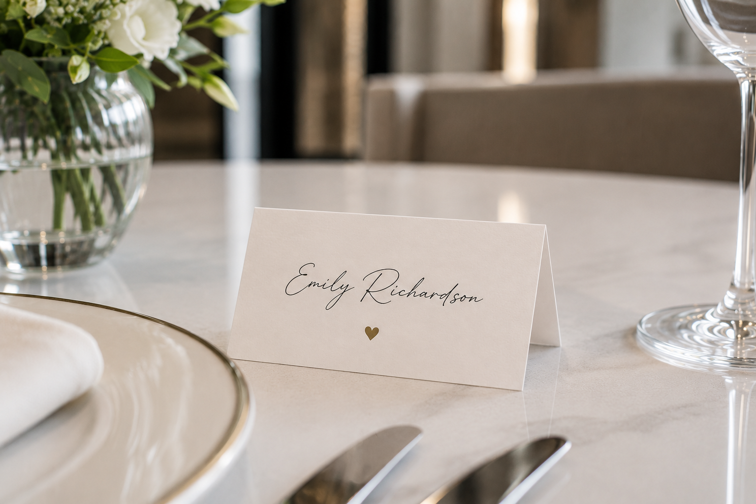 A place card folded on a table in front of a vase, for Emily Richardson. A small beige heart accompanies the calligraphic font used.